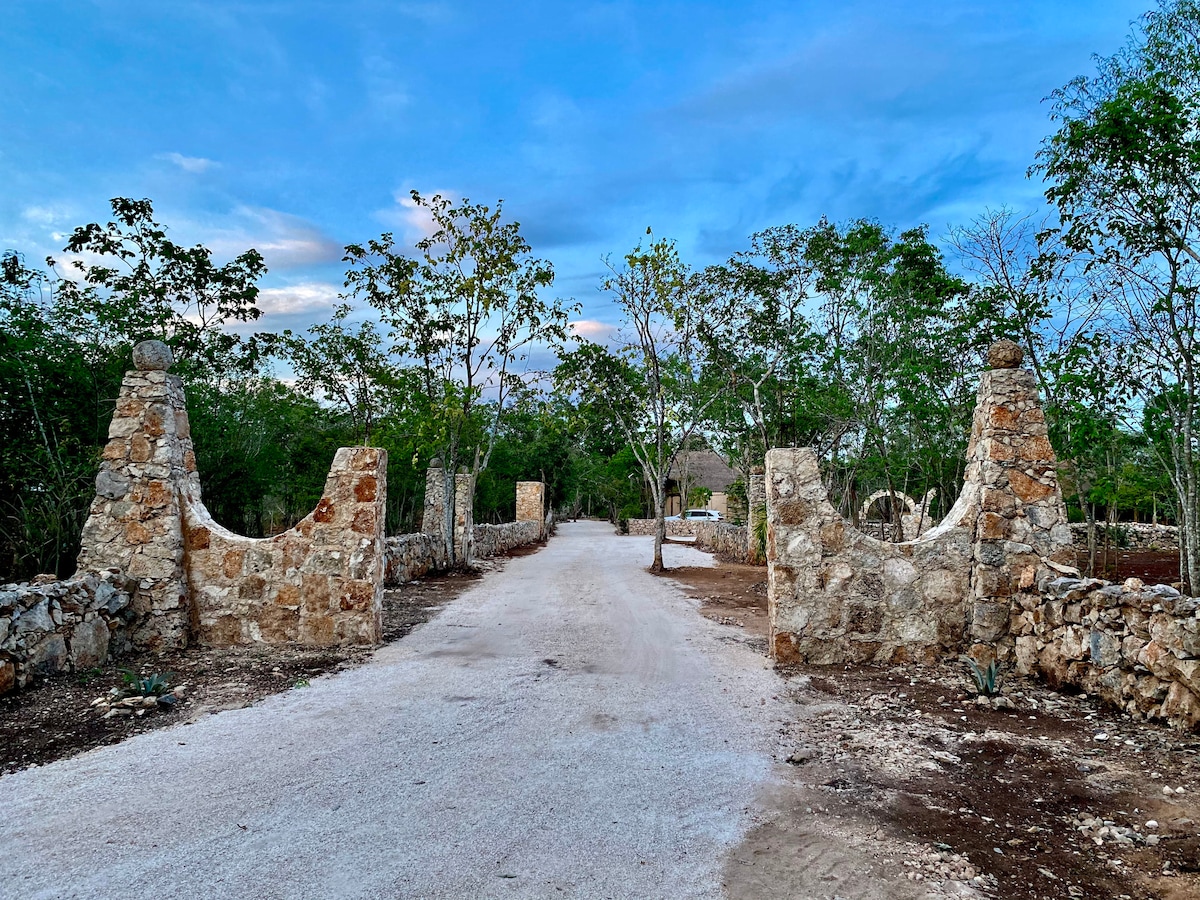 A rustic entrance is framed by stone pillars, leading to a winding path surrounded by greenery. A gravel road extends into the distance, flanked by low stone walls and clusters of trees, inviting a sense of exploration.