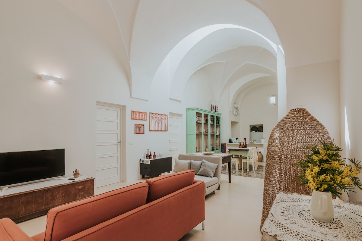 An open space living area is illuminated by natural light, featuring a cozy seating arrangement with orange sofas. A green shelving unit is positioned against the wall, accompanied by a dining table and chairs, while decorative elements add a welcoming touch to the room.