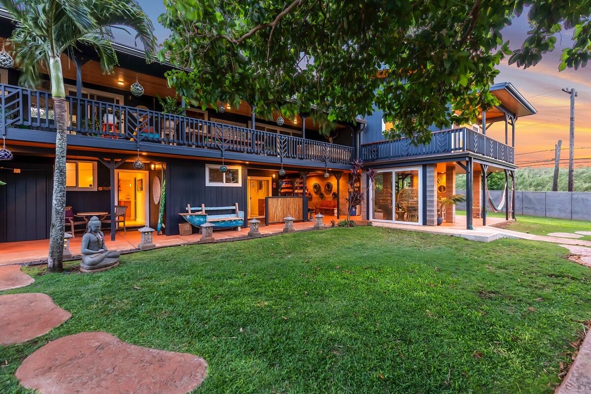 A spacious outdoor area is framed by lush greenery, featuring manicured lawns and pathways. The two-story structure presents a mix of dark exterior walls and welcoming balconies, with warm lighting visible through the windows. Decorative elements and a tranquil statue create a peaceful ambiance.