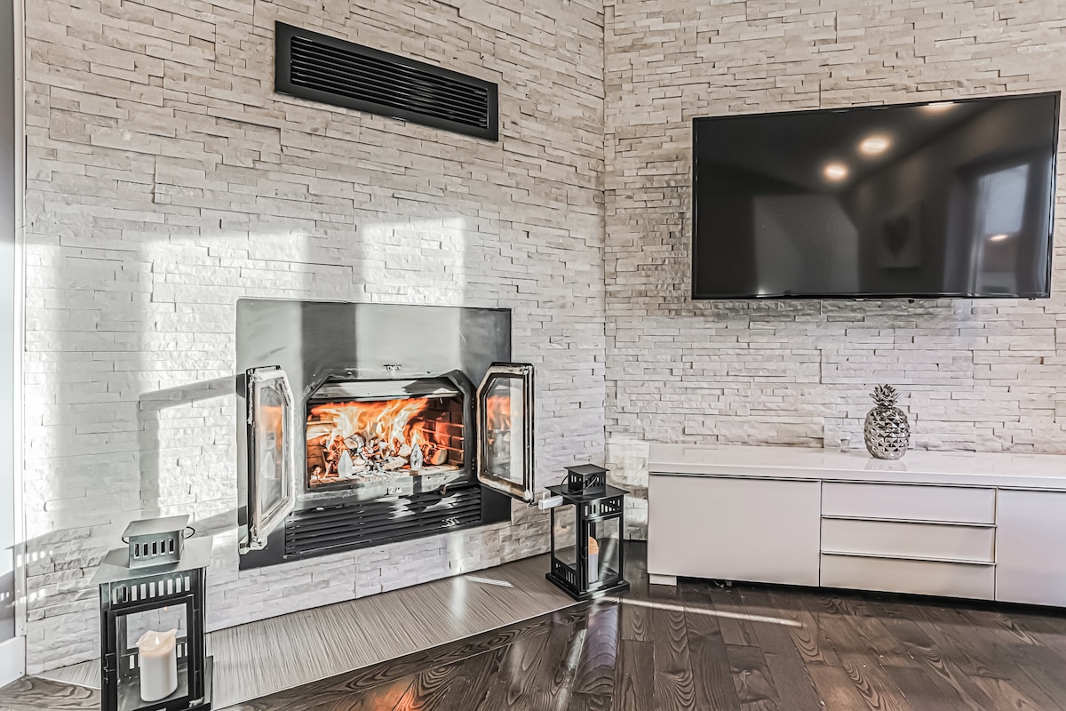 A modern fireplace is showcased within a textured stone wall, providing warmth and visual interest. Two lanterns, one with a candle, are placed nearby, complementing the sleek white media console beneath a mounted television.