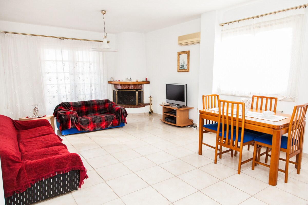 A spacious living area features a red sofa and a plaid-covered couch, complemented by a wooden dining table with seating for six. Light streams through large windows adorned with sheer curtains. A flat-screen TV is positioned near a decorative fireplace.