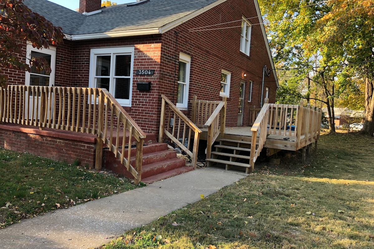 The exterior of a two-story brick duplex features a well-maintained yard with grassy areas. A wooden ramp and steps lead to the entrance, providing accessible access. Trees are visible in the background, adding greenery to the setting. Soft sunlight highlights the inviting atmosphere.