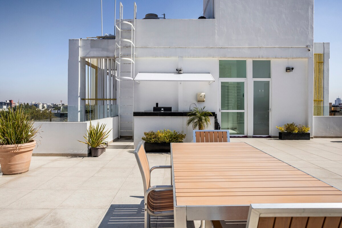 A rooftop garden is showcased with a large wooden table surrounded by several chairs. Potted plants are placed around the space, and a counter area is visible in the background. Clear skies create an open atmosphere in this elevated setting.