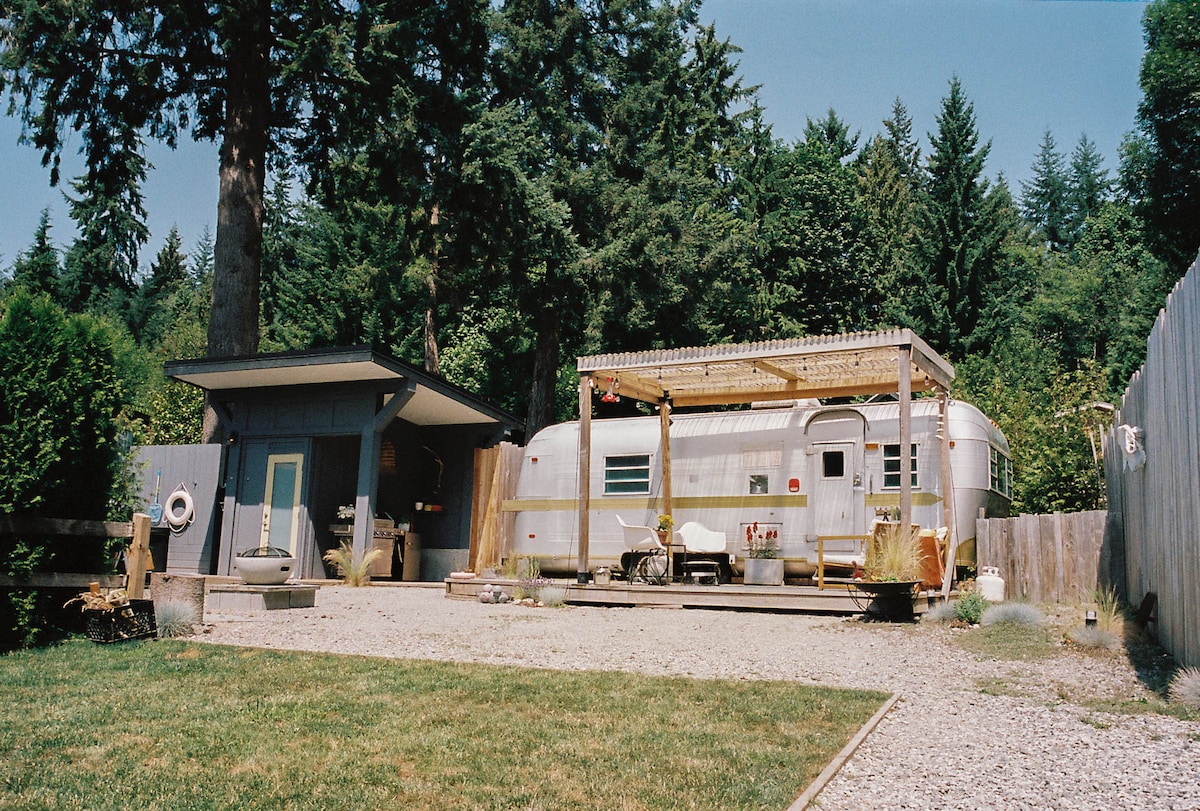 A renovated vintage trailer sits among lush greenery, with outdoor seating and a pergola providing shade. The well-maintained gravel area leads to a spacious lawn, and a detached structure is visible nearby, offering additional outdoor living space.