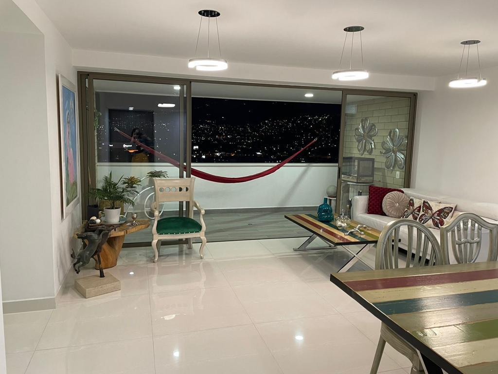 A spacious living area features a large window with a city view, surrounded by decorative elements. A hammock hangs in the background, and a dining table with colorful stripes is positioned alongside comfortable seating. Indoor plants and artwork add to the environment.