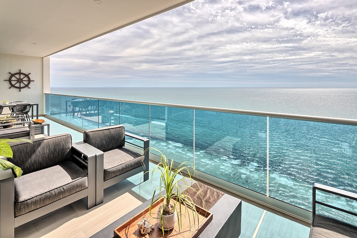 A modern balcony area offers comfortable seating with gray couches and a coffee table, overlooking the expansive Sea of Cortez. The glass railing provides an unobstructed view of the water and sky, creating a serene outdoor retreat.