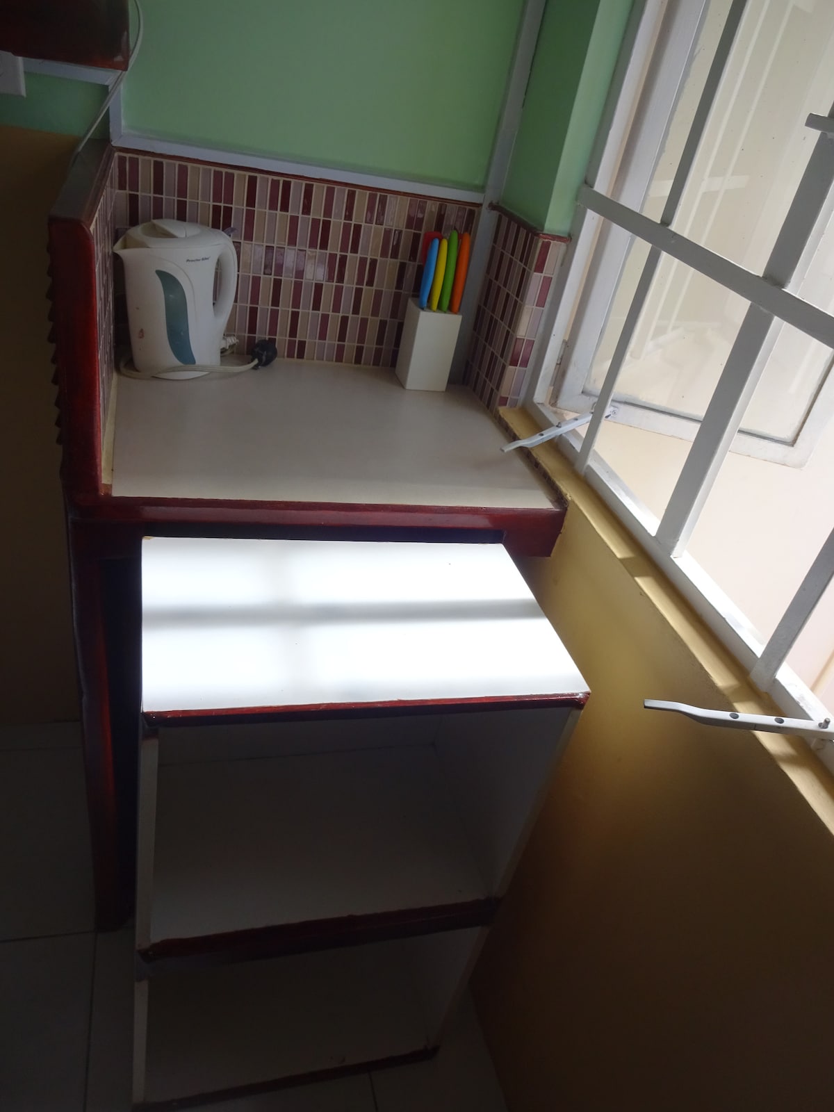 A compact kitchenette corner features a small countertop with a kettle and a holder for colored utensils. Natural light filters through a window, illuminating the space. A minimalistic storage unit sits beneath the countertop, enhancing functionality.