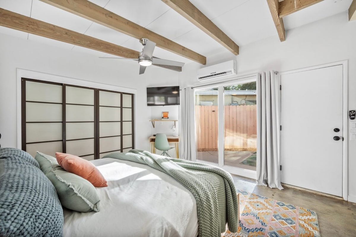 A spacious bedroom is highlighted by natural light streaming through sliding glass doors. A California King bed is adorned with soft pillows and a textured blanket. Exposed wood beams add character to the ceiling, while a workspace is positioned near the entrance.