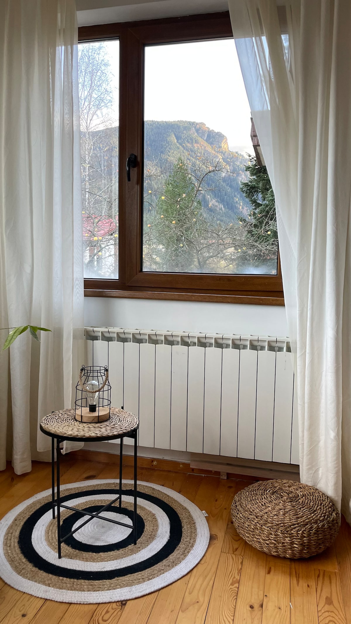 A cozy corner features a large window offering mountain views, framed by sheer white curtains. A round black and white rug lies beneath a small, round table with a lantern. A woven pouf complements the natural wood flooring and warms the inviting space.