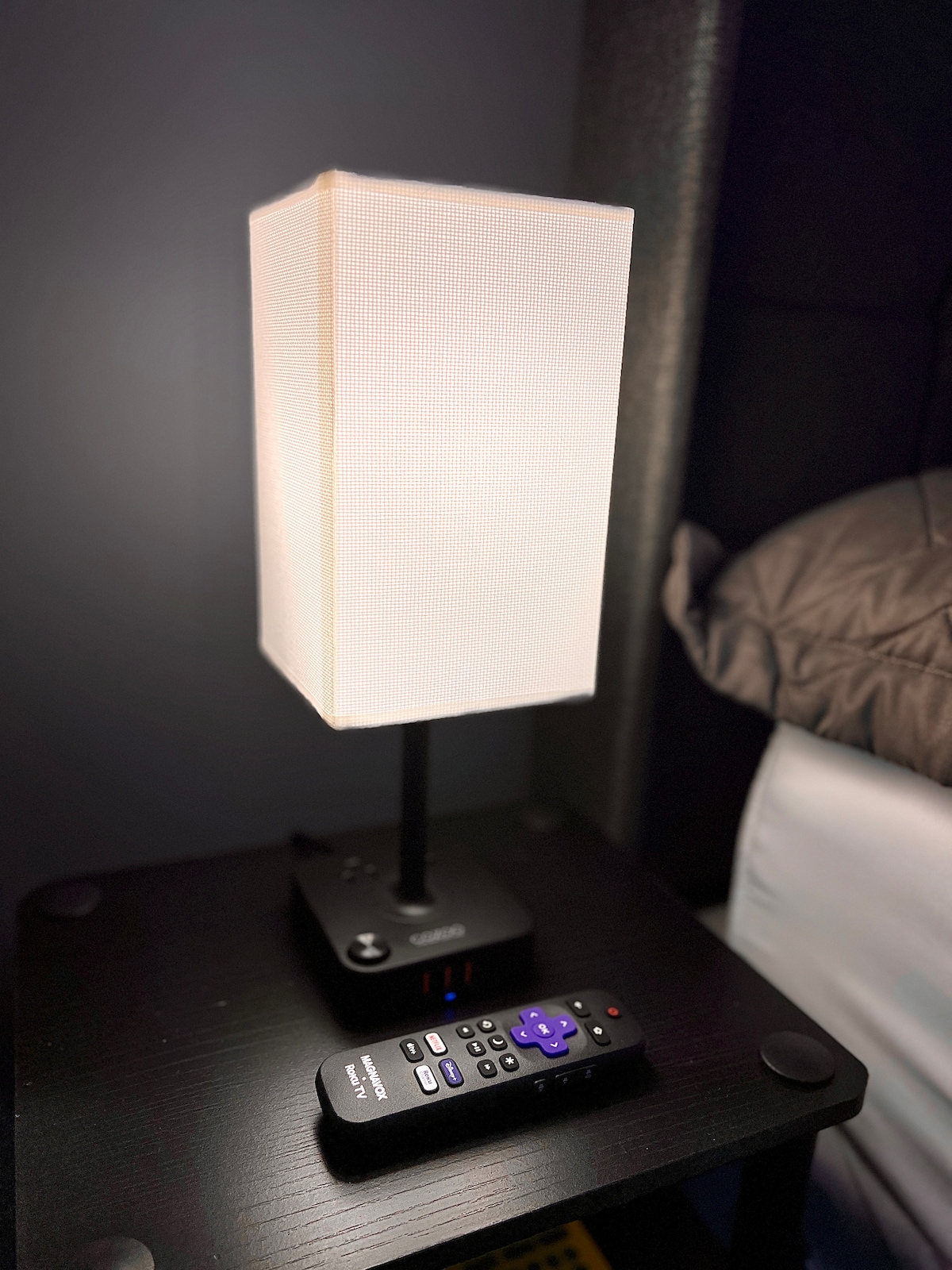 A bedside table is equipped with a modern lamp featuring a textured shade. Next to the lamp, a remote control lies on the dark surface, indicating ease of access for guests. The background shows part of a bed with muted bedding.