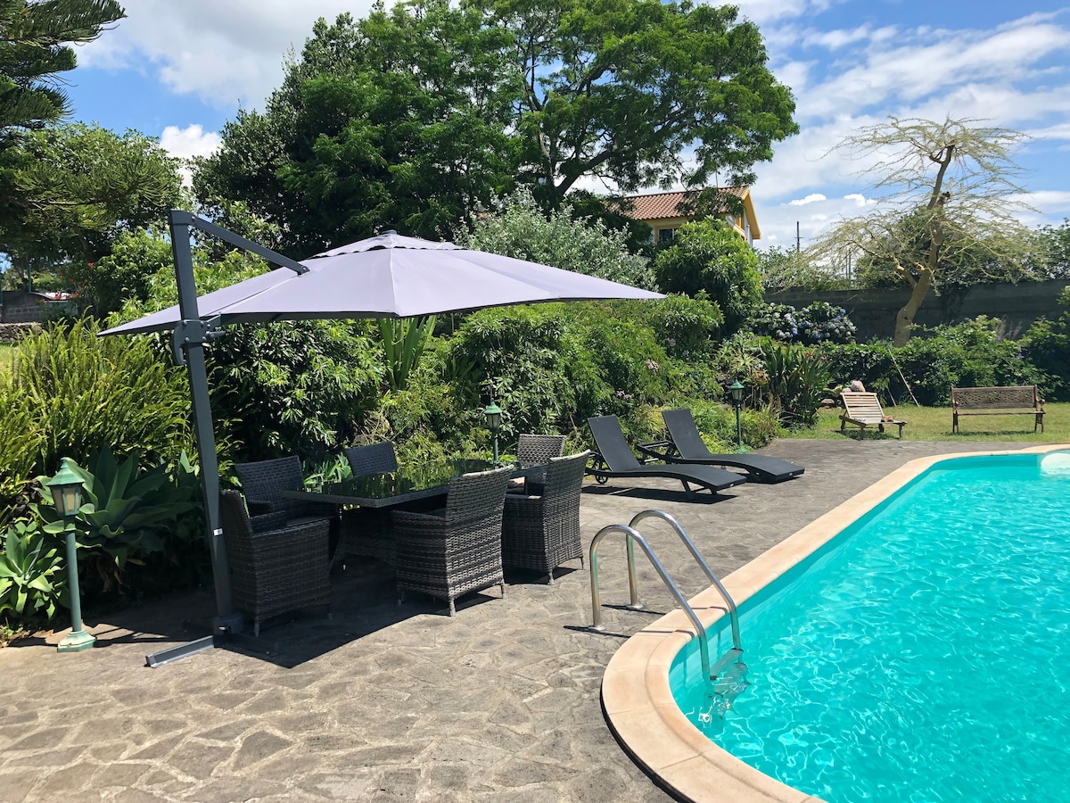 A pool area features a light blue swimming pool bordered by large natural stone tiles. Comfortable lounge chairs are positioned nearby, along with a shaded dining set under a large umbrella. Lush greenery and trees surround the space, contributing to a serene atmosphere.