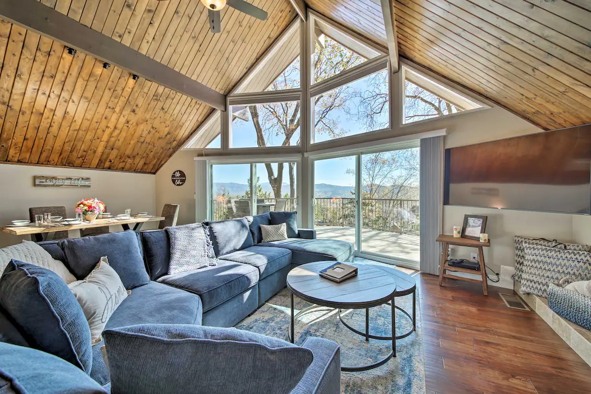 A spacious living area features a large sectional sofa in soft blue, complemented by wooden ceilings and expansive windows that fill the room with natural light. A round coffee table is positioned on a plush area rug, with views of the outdoors visible through the glass walls.