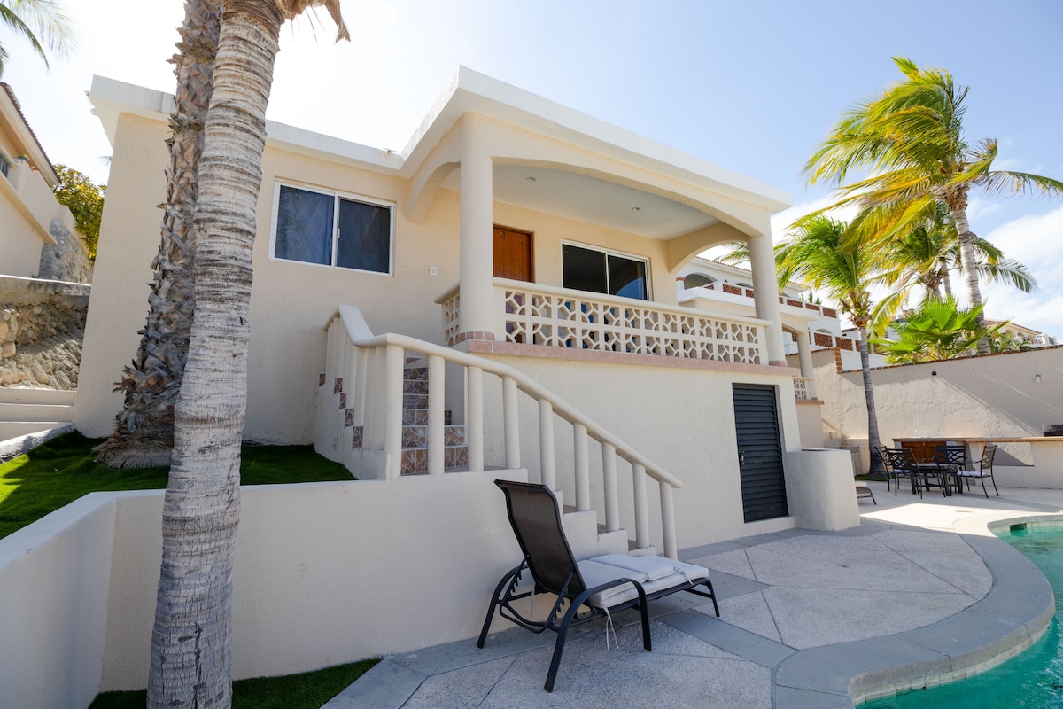The exterior of the villa is showcased, featuring a light-colored stucco facade and a curved balcony. Palm trees are visible alongside the property, adding a tropical feel. A staircase leads to the entrance, and outdoor seating is arranged near the pool area.