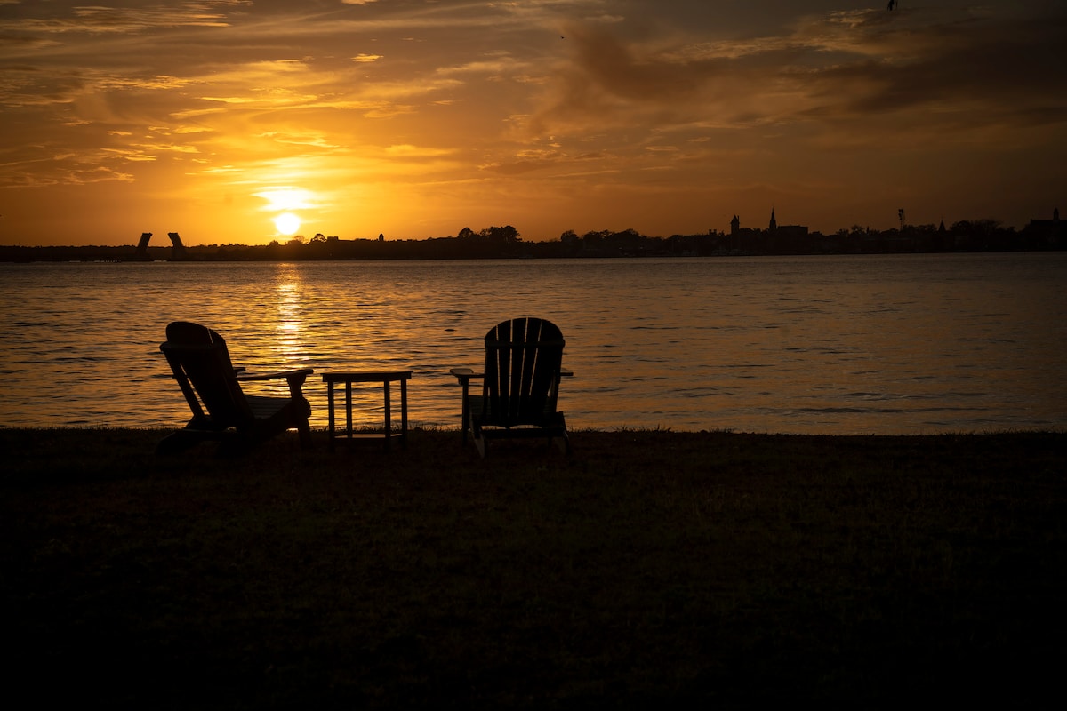 Two Adirondack chairs and a small table are positioned on a grassy area near the water's edge, facing a tranquil sunset over the Neuse River. Silhouetted against the glowing horizon, the scene is serene, inviting relaxation and contemplation.