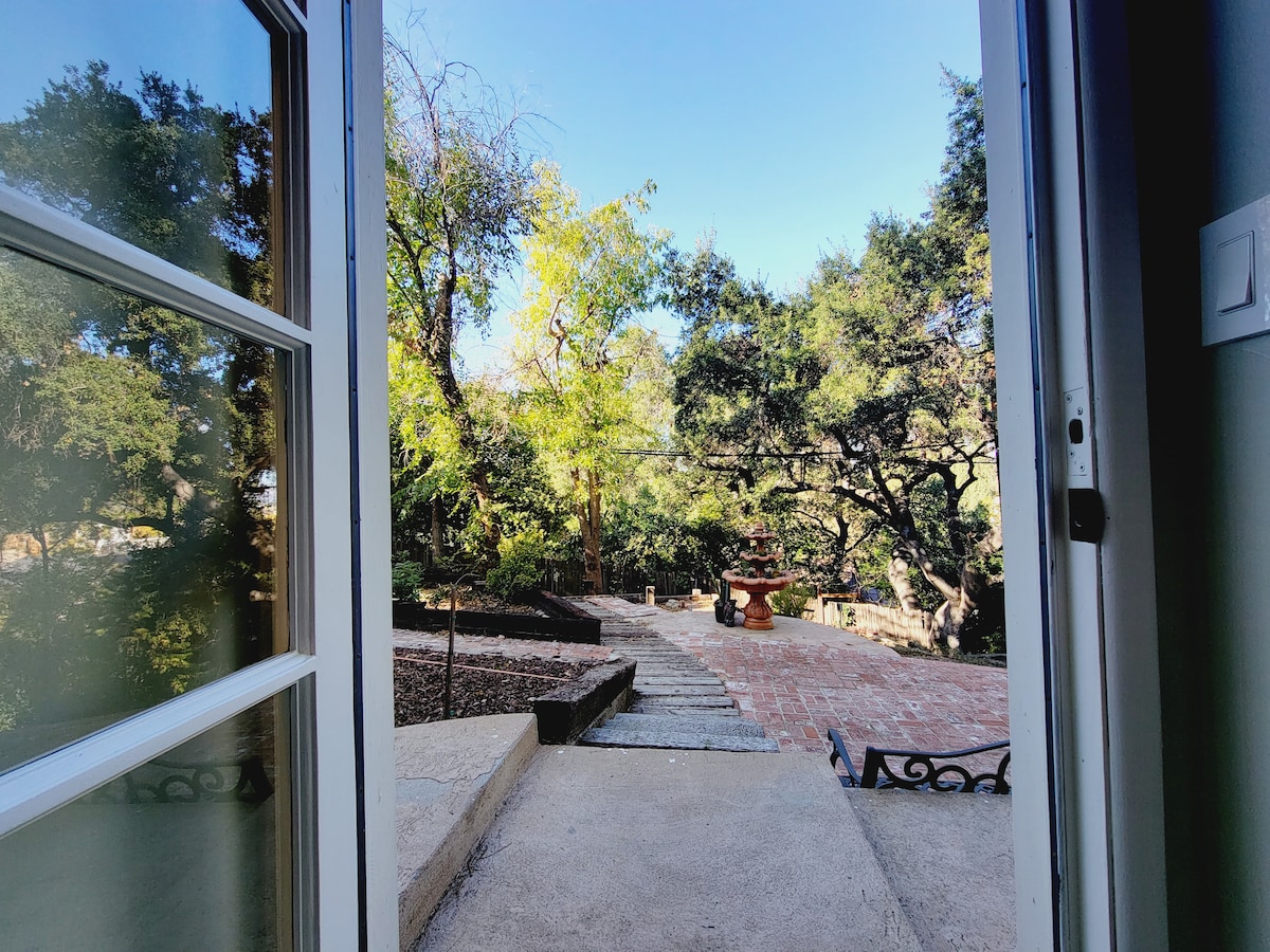 An open door reveals a private backyard garden, featuring stone steps leading to a patio area surrounded by lush trees. Natural light filters in, creating a connection between indoor and outdoor spaces. A serene atmosphere and potential for relaxation are emphasized.