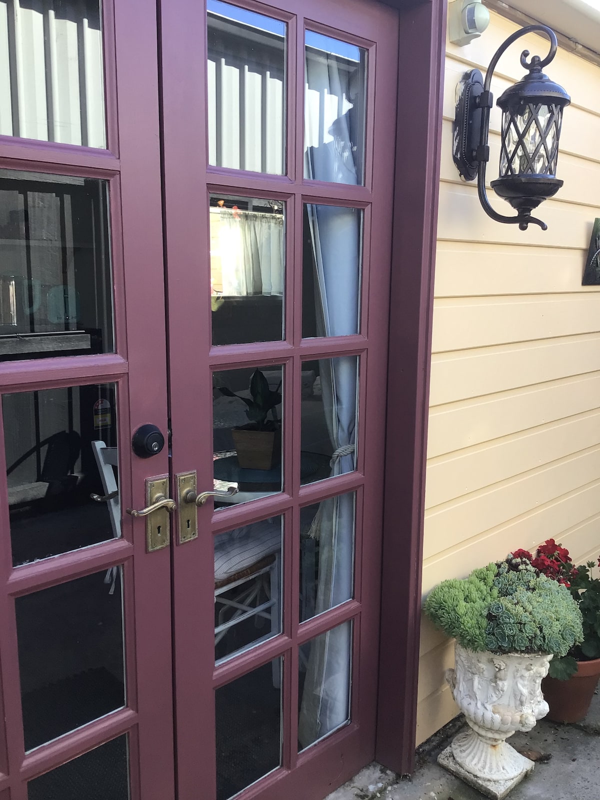 A set of double doors with multiple glass panes is framed by a warm-colored exterior wall. A decorative lantern is mounted beside the entrance, and a potted plant with vibrant flowers adds a touch of greenery to the scene.