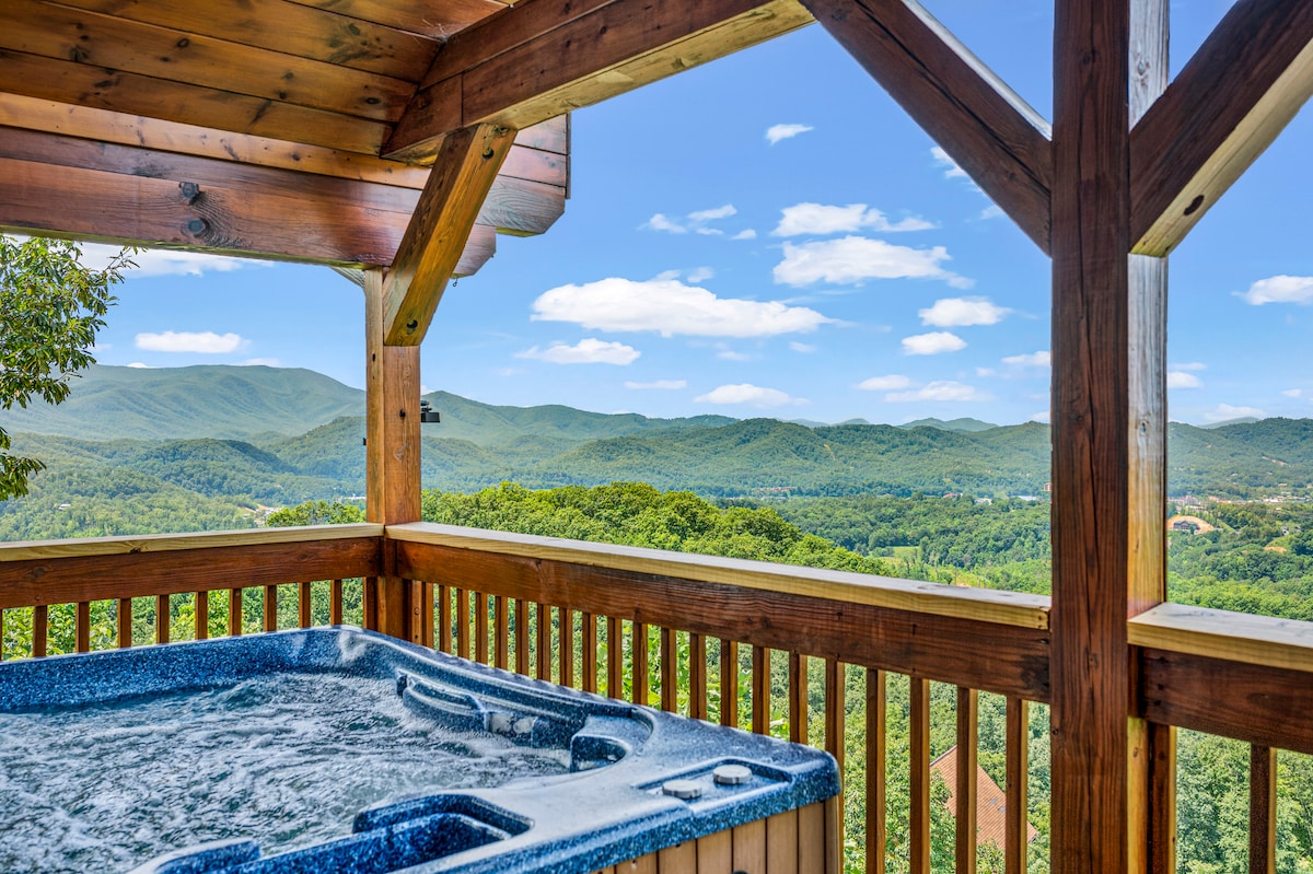 A hot tub is positioned on a wooden deck, featuring views of lush green mountains under a clear blue sky. The surrounding railing showcases the expansive scenery, enhancing the connection to the tranquil, natural environment.