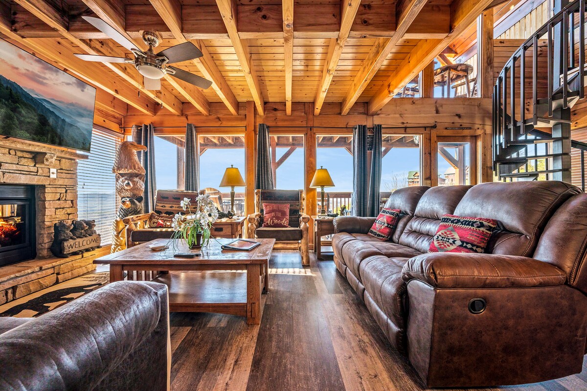 A spacious living room showcases wooden beams and large windows, enhancing natural light. Comfortable leather sofas are arranged around a coffee table, with decorative pillows adding warmth. A stone fireplace is visible, providing a cozy focal point. Scenic mountain views are framed by the windows.