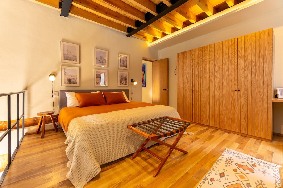A spacious bedroom features warm wooden beams across the ceiling. A large bed with an orange duvet is accompanied by matching decorative pillows. A natural wood wardrobe and a simple wooden side table add to the minimalist decor, while a patterned rug is laid on the wooden floor.