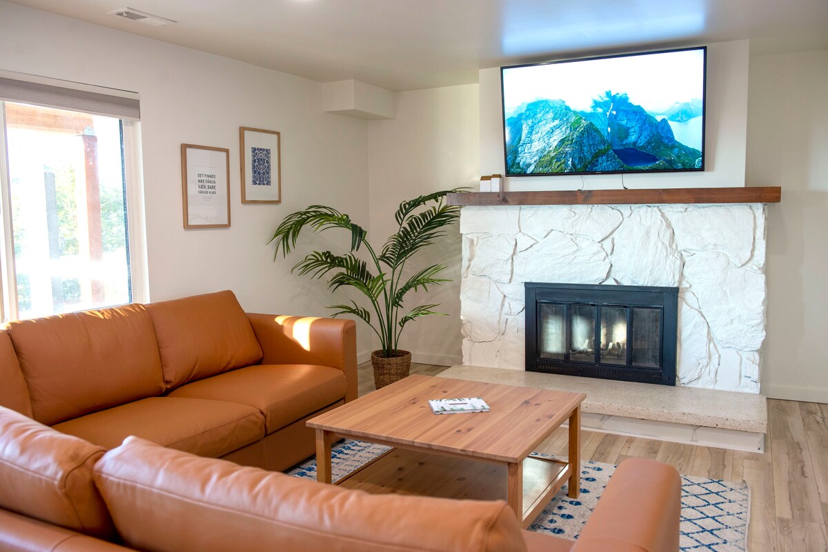 A living area features a stone fireplace with a mounted television above. Two brown leather sofas are arranged around a wooden coffee table on a patterned rug. A potted plant adds a touch of greenery, while natural light enters through a window.
