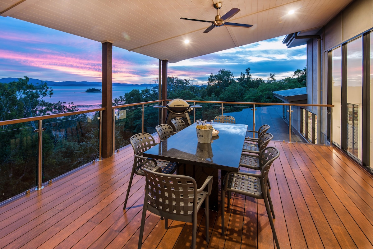 An outdoor dining area is set on a wooden deck, featuring a large table surrounded by nine black chairs. A barbecue grill is visible to the side, with panoramic views of the surrounding landscape and a pastel sunset in the background.