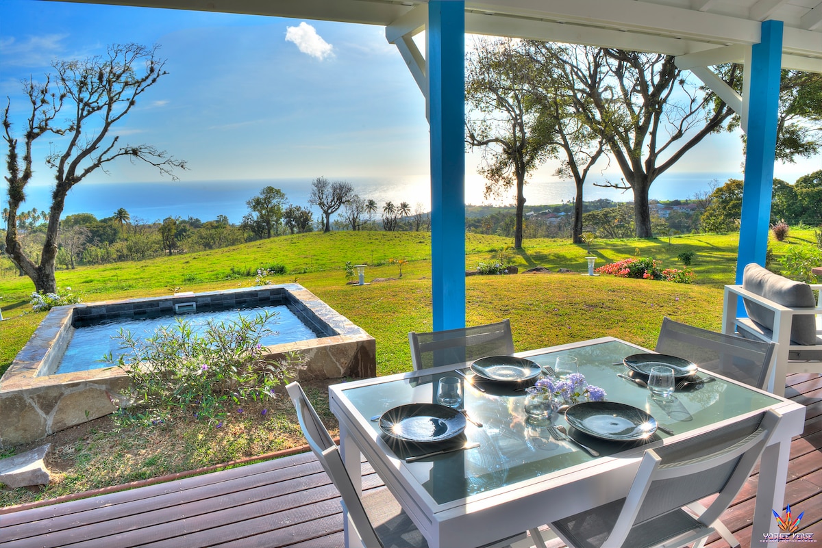 A dining area is set outside under a covered patio, featuring a glass table with plates and glasses arranged on top. In the background, a private soaking tub can be seen amidst the lush greenery, leading to views of the Caribbean Sea and the horizon.