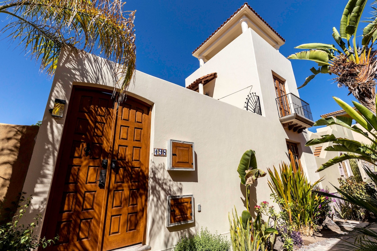 A charming two-story house is presented, featuring a warm wooden door framed by lush tropical foliage. The building's white exterior contrasts against the vibrant blue sky, while a balcony extends above the entrance, enhancing the architectural appeal.