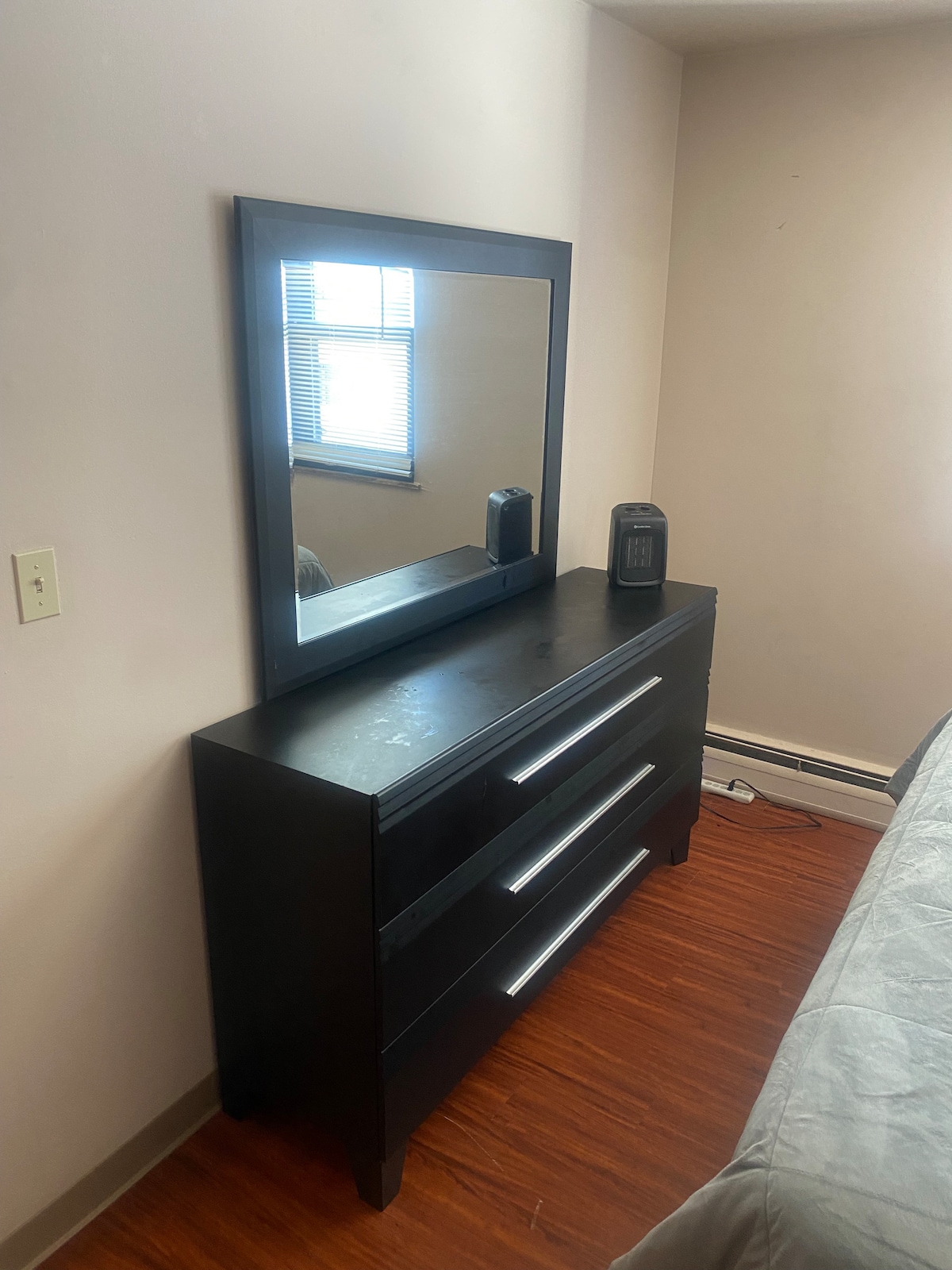 A black dresser is positioned against a light-colored wall, featuring three spacious drawers with sleek, metal handles. Above the dresser, a rectangular mirror reflects natural light coming from a nearby window. An electric heater is placed next to the dresser.