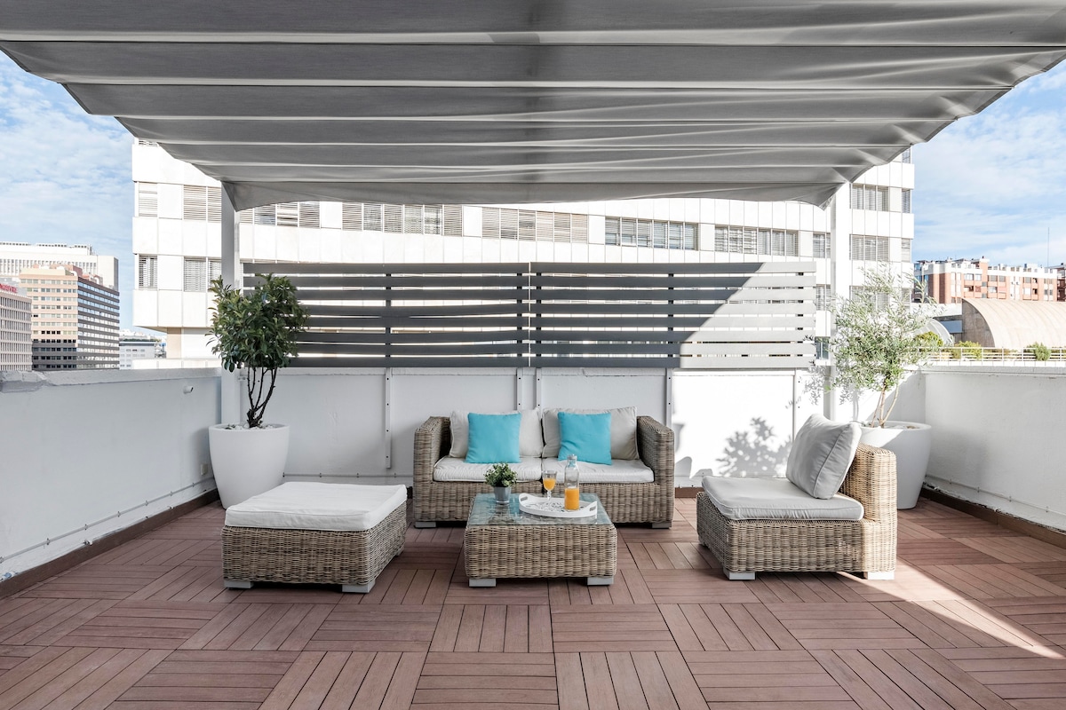 A spacious terrace is highlighted with modern outdoor furniture including two lounge chairs and a coffee table. Potted plants add greenery, while light fabric canopies provide shade. The wooden flooring complements the urban backdrop, featuring city buildings visible in the distance.