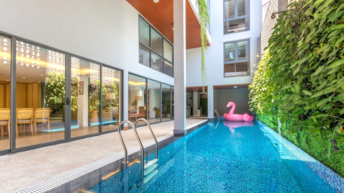 An inviting swimming pool is flanked by modern architecture and lush greenery. Glass walls enhance the illusion of space, while a vibrant pink float adds a playful touch to the tranquil blue waters. Comfortable seating areas can be glimpsed through the large glass doors.