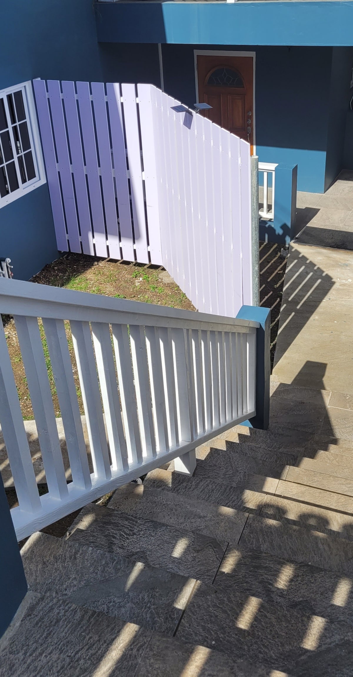 A staircase leads down to an outdoor area, framed by white railings and a light purple fence. The path is made of stone, and a wooden door is partially visible at the top of the staircase. Sunlight casts soft shadows on the steps.