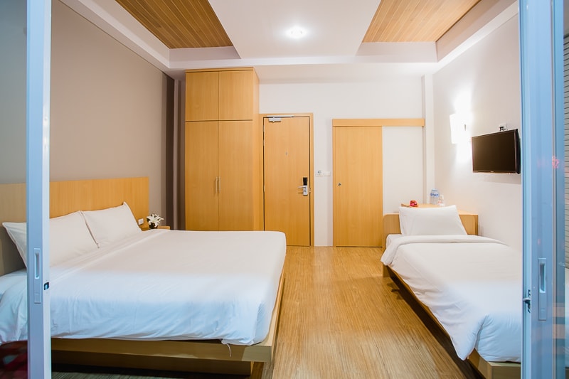 A well-organized room is presented with wooden accents and neutral tones. Two beds are positioned, one larger and another single, with white bedding. A television is mounted on the wall opposite the beds. The entrance door and a closet are visible, enhancing overall accessibility.
