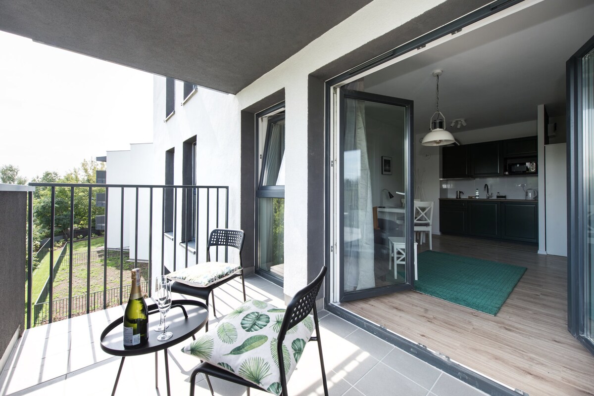 A balcony is featured, furnished with two black chairs and a small table. Open glass doors reveal a glimpse of the kitchen area, while greenery is seen in the background, suggesting a tranquil setting.