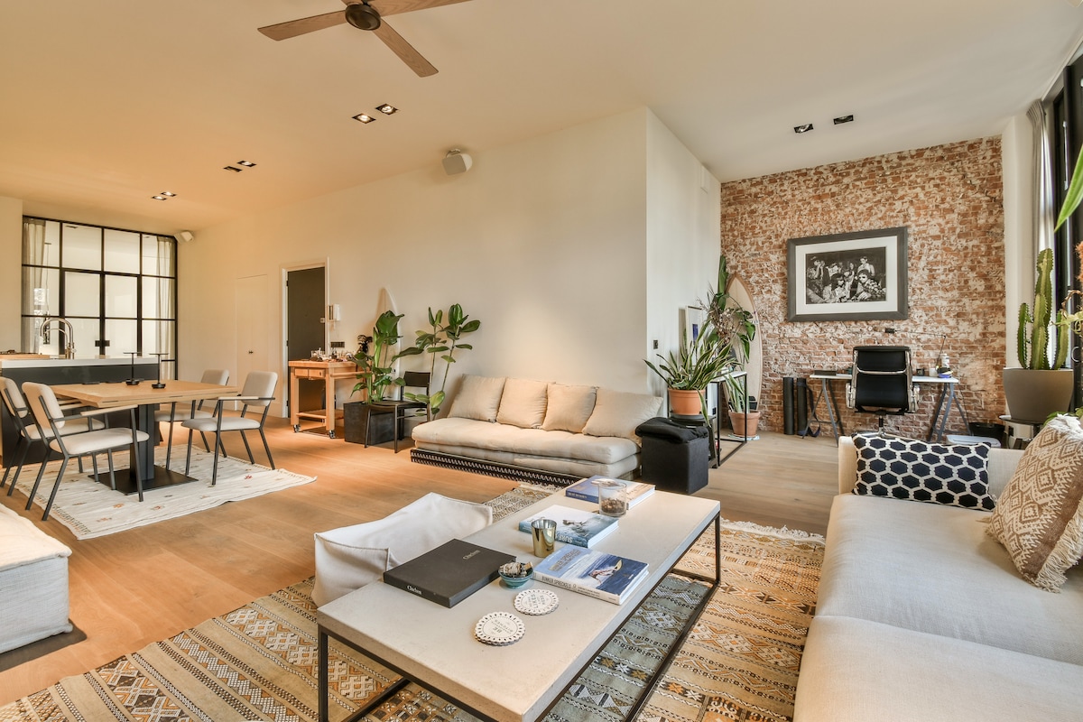 A spacious living area features a mix of modern design furniture and greenery. A comfortable seating arrangement includes a large sofa and stylish armchairs, complemented by a dining table that seats six. Exposed brick walls and large windows enhance the open ambiance.