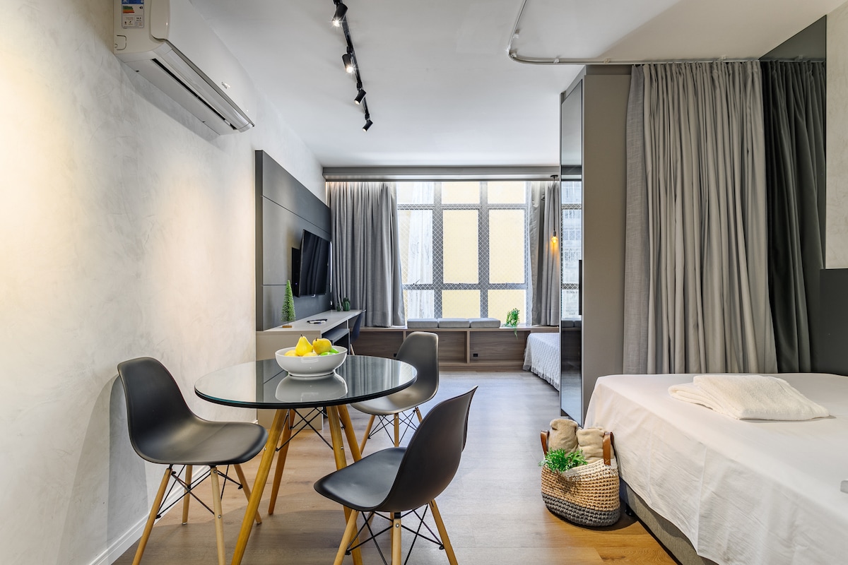 A modern studio apartment is highlighted by a round dining table with four black chairs. Soft curtains frame large windows, allowing natural light to fill the space. A comfortable bed is partially visible, offering a cozy atmosphere in the open layout.