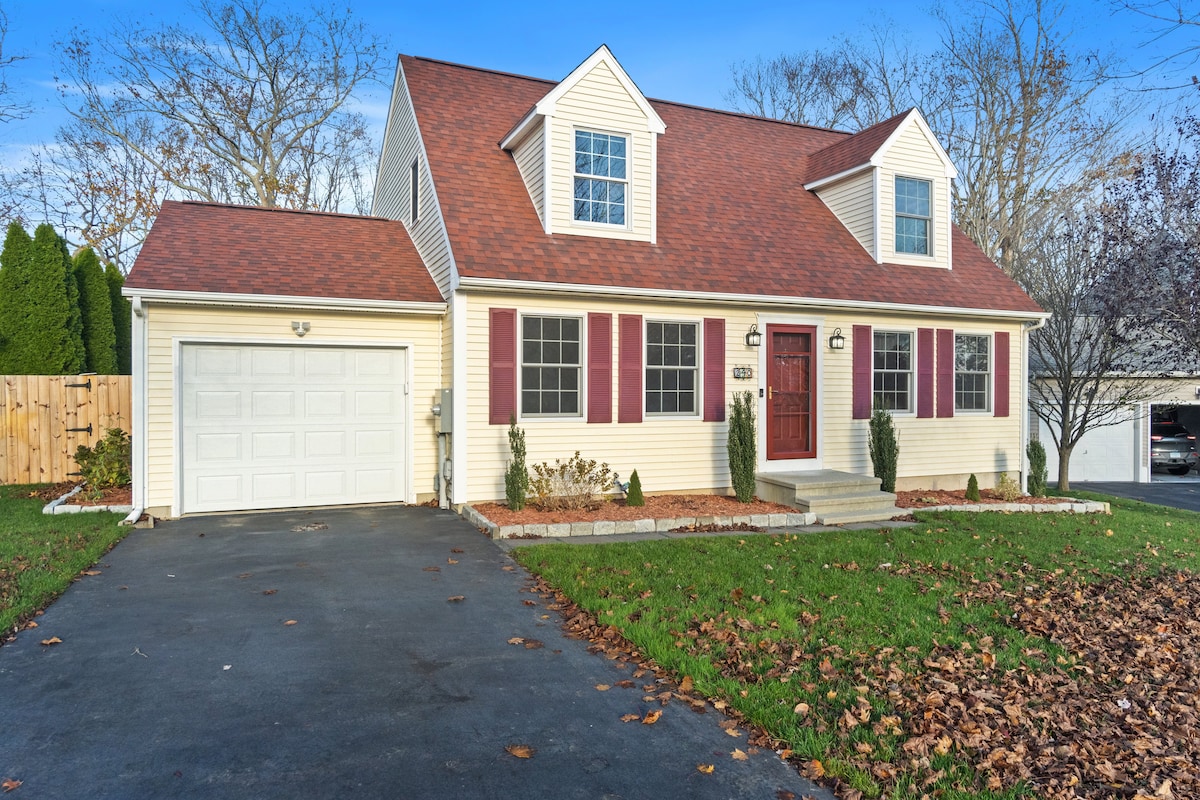 A two-story house features a welcoming front porch with a red door. Symmetrical windows are adorned with maroon shutters, while landscaping includes shrubs bordering the entrance. A driveway leads to a garage, and fallen leaves cover the lawn, signifying the autumn season.