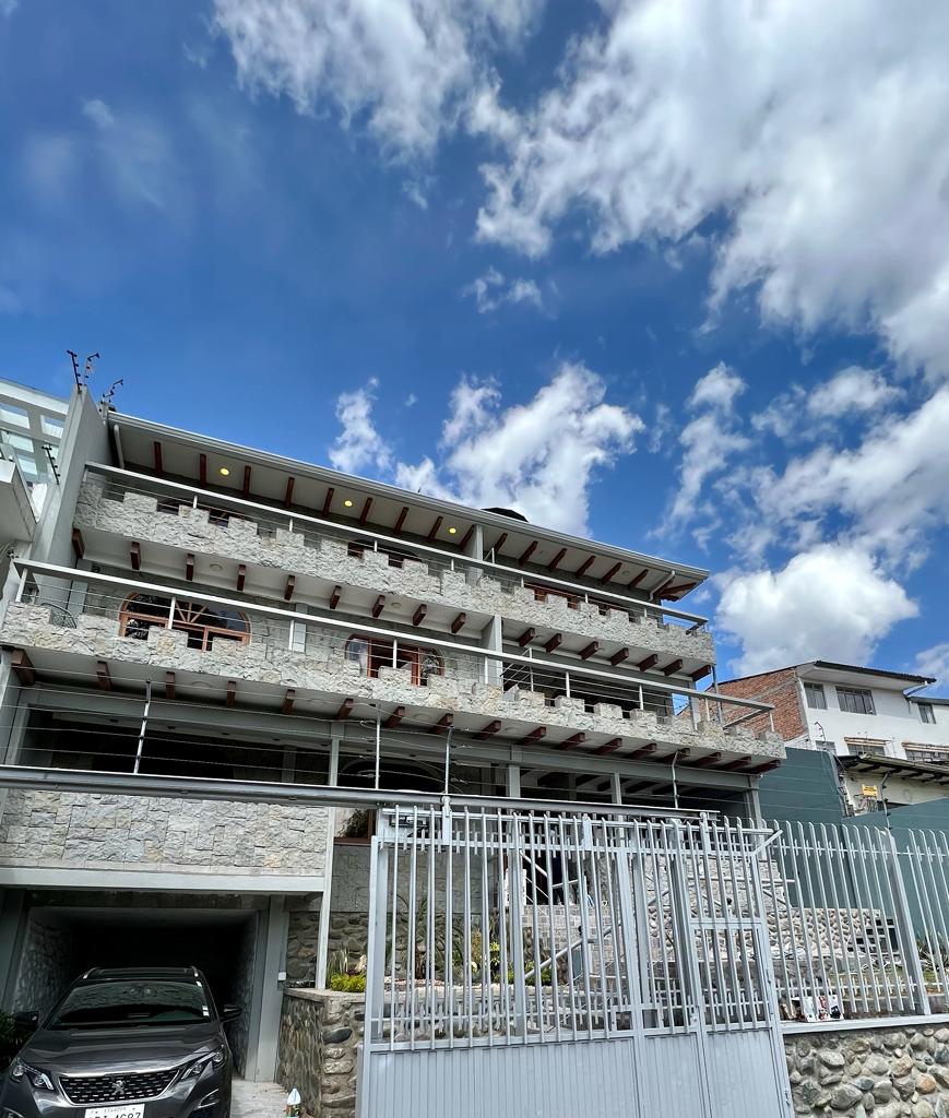 A modern multi-story building is presented, featuring a combination of stone and wood elements. Balconies with wooden railings extend from each floor, allowing for outdoor seating. A clear blue sky with scattered clouds serves as the backdrop, enhancing the building's contemporary design.