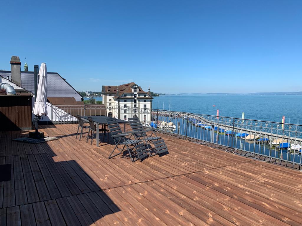 A large wooden deck area features outdoor seating, including a dining table and lounge chairs. The view overlooks a tranquil lake with boats visible on the water. A railing provides safety while maximizing the scenic lakefront experience.