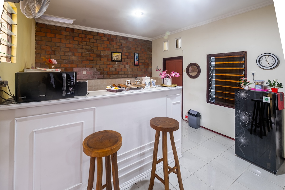 A compact kitchen area is showcased, featuring a wooden breakfast bar with two stools. The brick wall adds a rustic touch, while modern appliances such as a microwave and a refrigerator are visible. Light tiles reflect natural light, contributing to a clean and functional space.