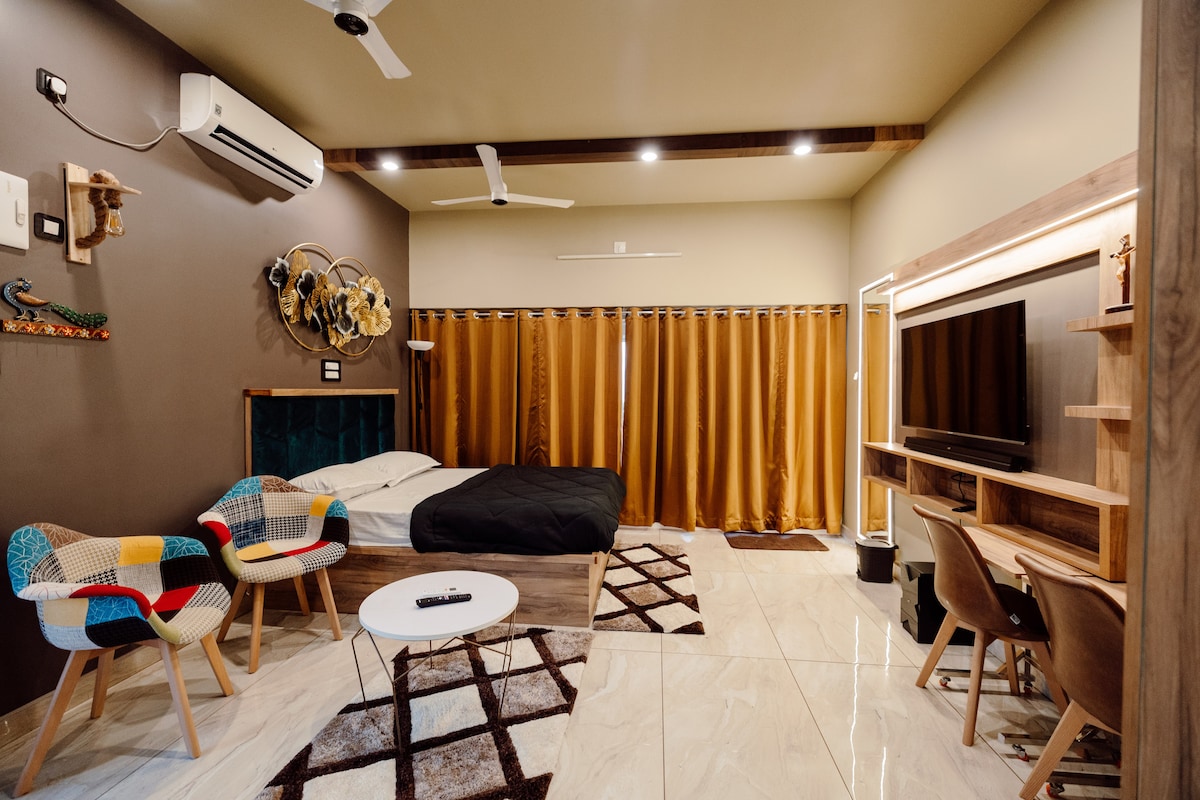 The studio apartment features a queen-size bed, a cozy seating area with colorful chairs, and a wooden TV unit with a wall-mounted LED TV. Large curtains partially cover the windows, and a decorative wall piece adds interest to the space.