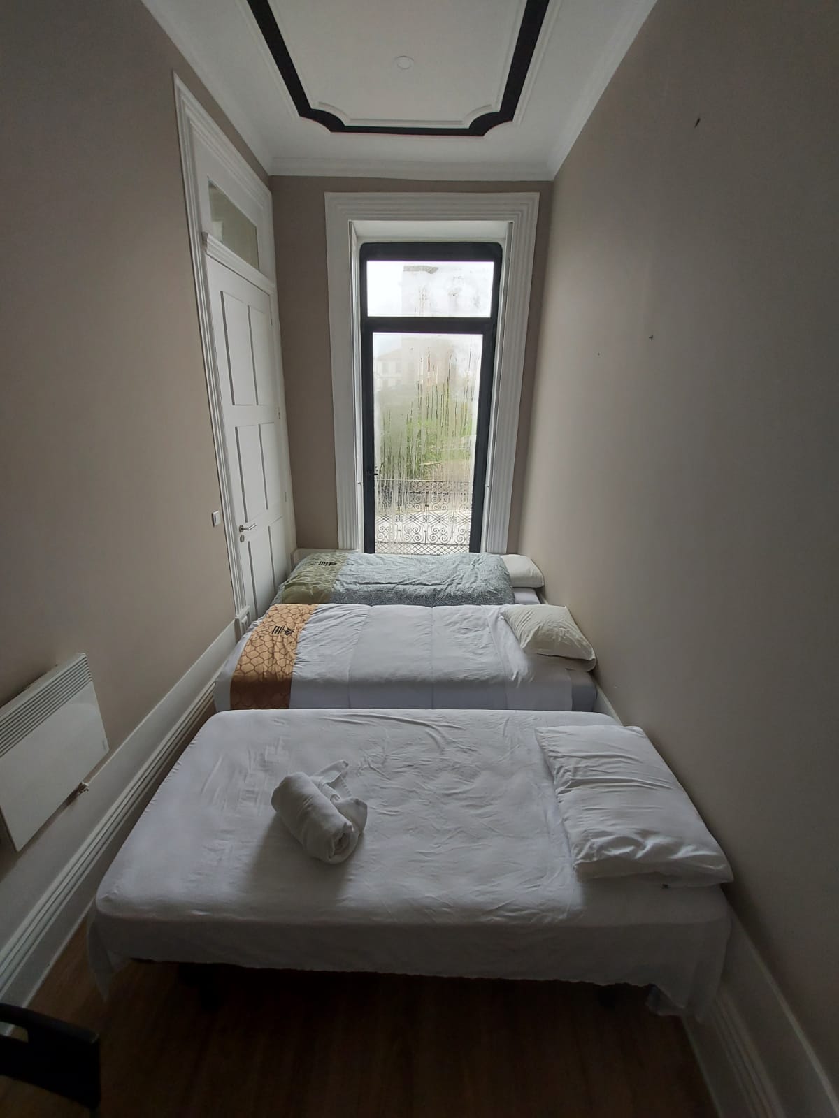 A cozy guest room is shown, featuring two single beds neatly arranged with white linens. A window allows natural light to enter, displaying a view of greenery outside. A towel is placed on one of the beds, and ample floor space is available.
