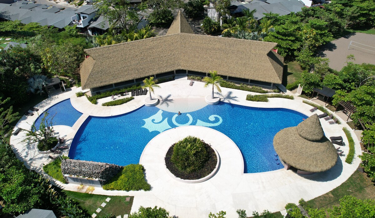An aerial view of a large swimming pool surrounded by well-maintained gardens. The pool features a unique circular design with shaded seating areas, palm trees, and a thatched-roof pavilion nearby, creating a serene environment for relaxation and leisure.