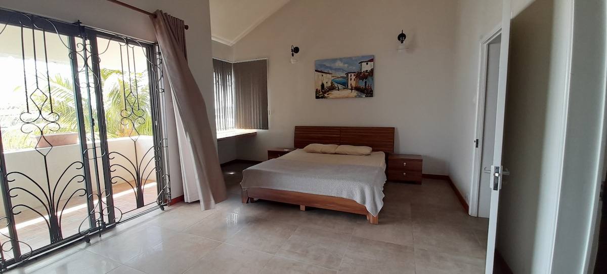 A spacious bedroom features a large bed with a neutral-toned blanket. Natural light enters through large windows and a glass door, which offers access to a balcony. The room is accented by light-colored walls and a piece of artwork depicting a serene landscape.