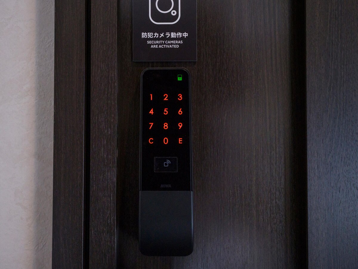 A door lock keypad is displayed, featuring illuminated red numbers and a security camera notice above. The keypad is mounted on a dark wooden door, with a green light indicating readiness. A symbol for accessibility is located at the bottom.