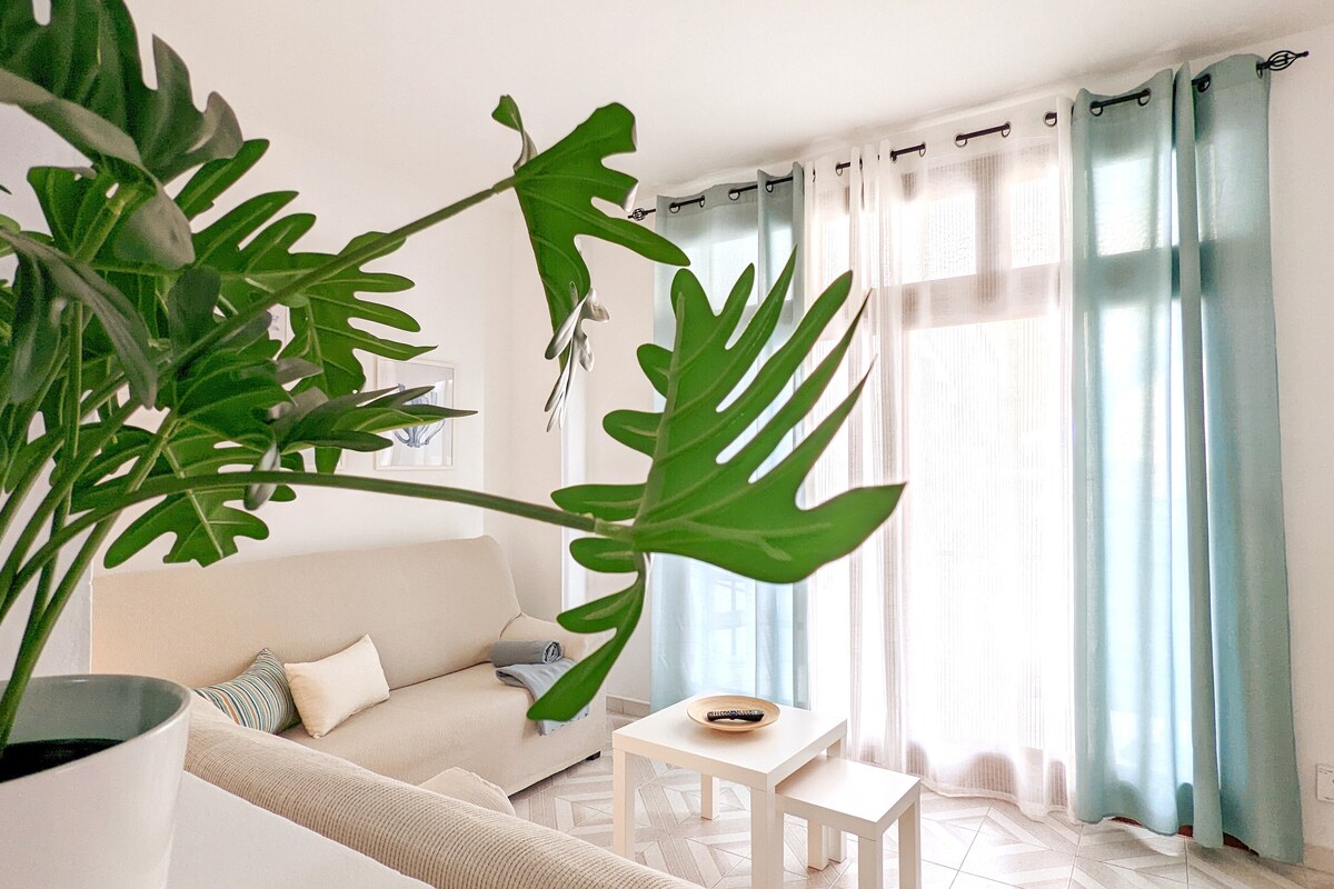 A bright living area is visible, featuring light-colored sofas with a small table in the center. Sheer curtains frame large windows, allowing natural light to fill the space. A leafy plant adds a touch of greenery to the scene, enhancing the room's overall freshness.