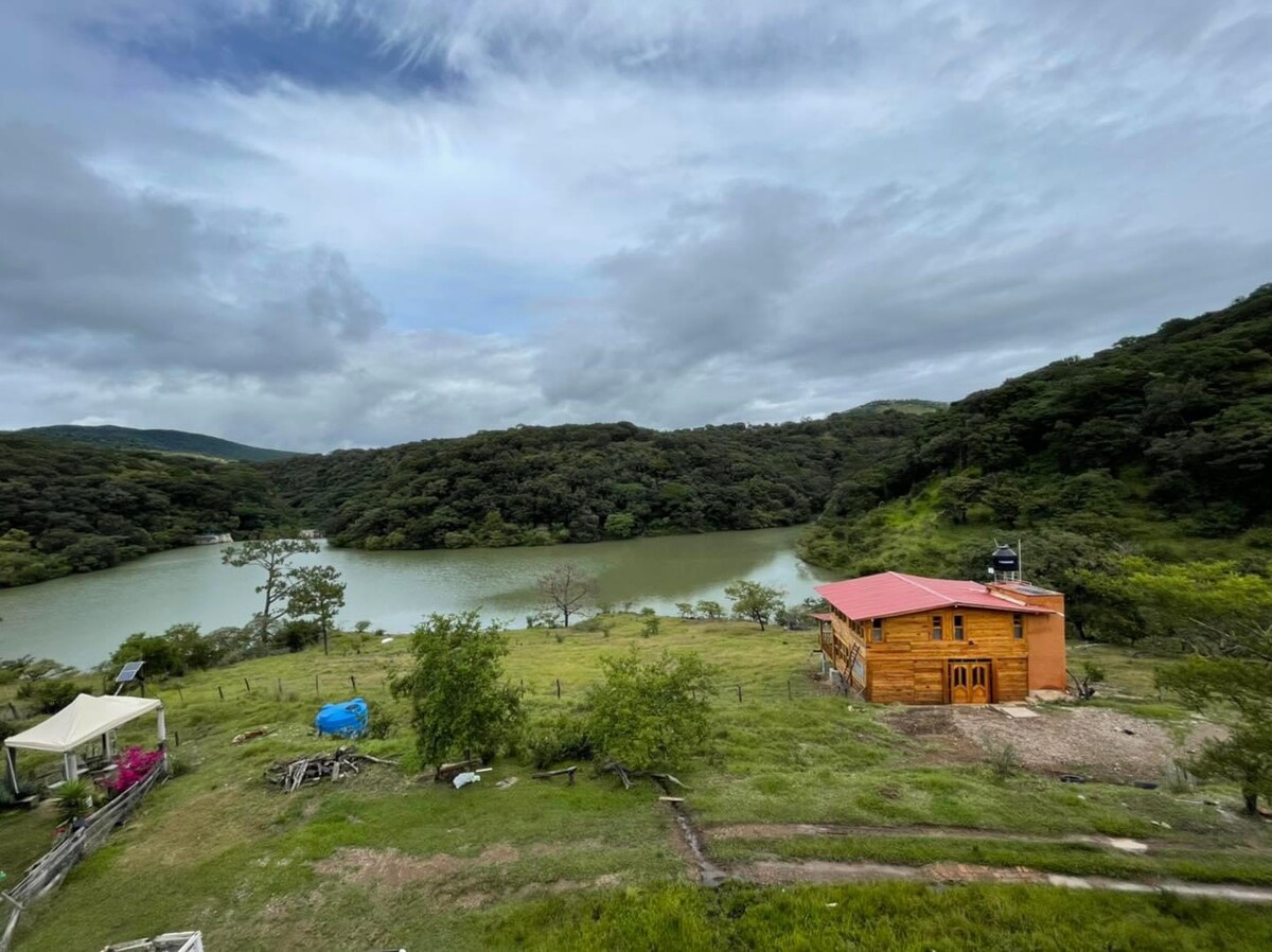A wooden cabin with a red roof is positioned near a calm lake, surrounded by lush greenery and rolling hills. The landscape features patches of grass and scattered trees under a cloudy sky, depicting a serene and natural environment.