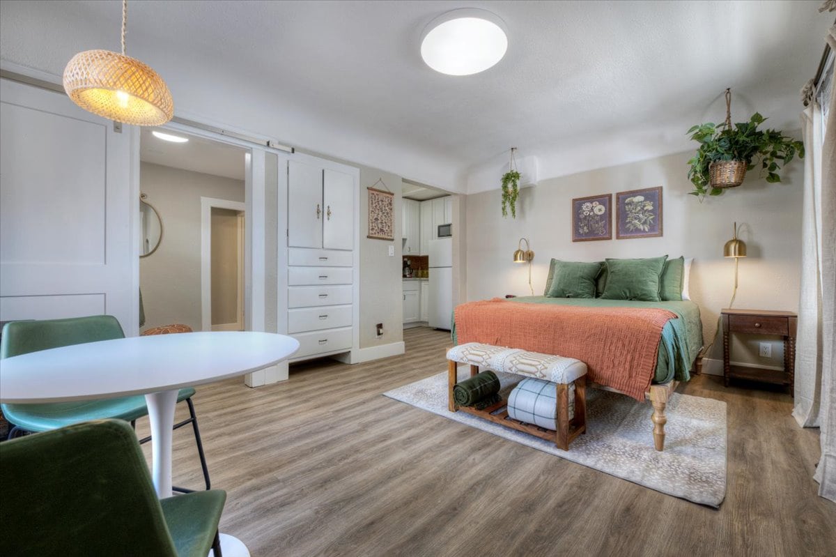 A cozy main living area is designed as a multifunctional space with a spacious bed adorned in earthy tones. A small dining table with green chairs complements the warm wood flooring. Natural light illuminates the room through a circular ceiling light. Decorative plants and artwork add character.