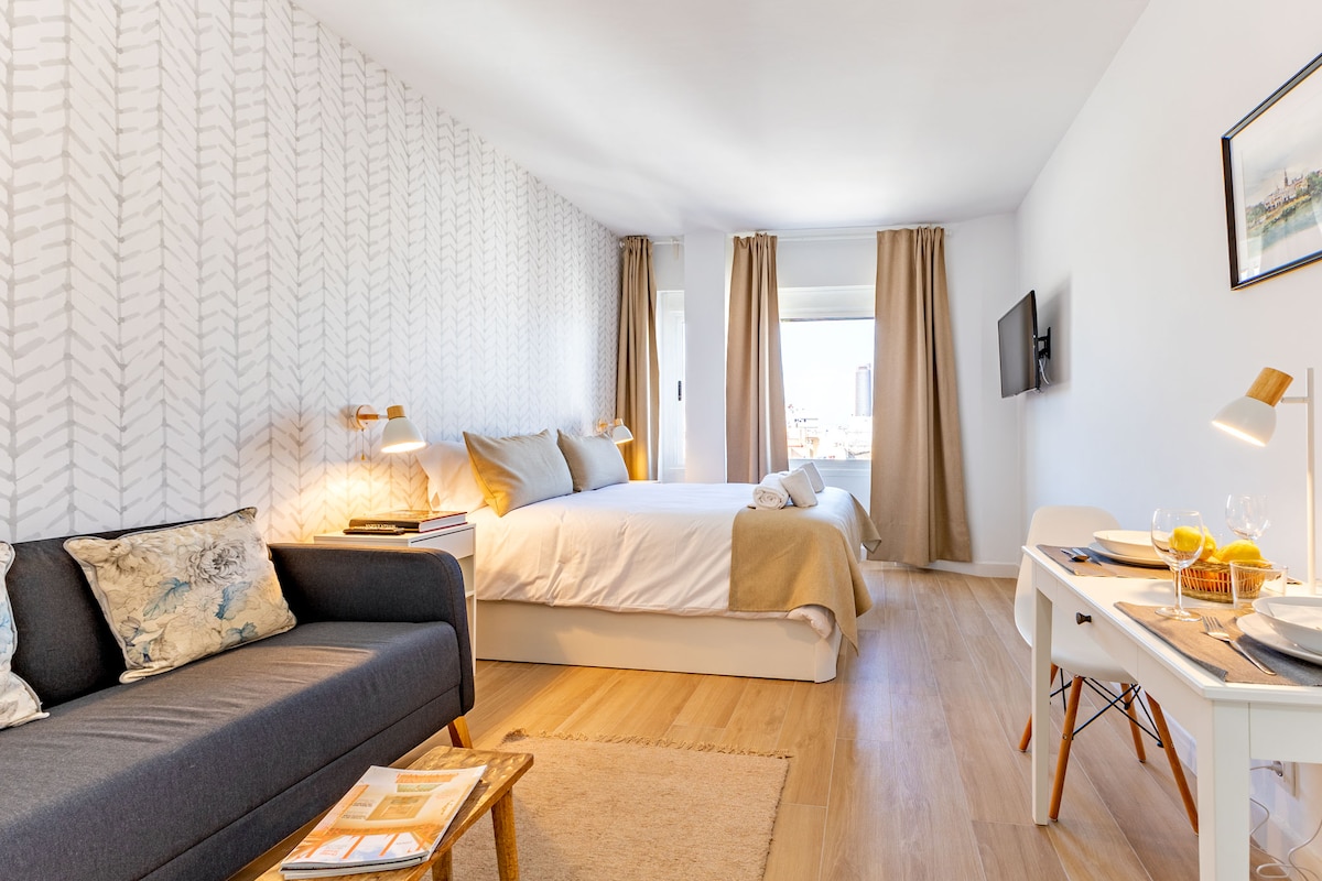 A well-lit room features a spacious bed dressed in neutral tones, complemented by light curtains. A cozy sofa is positioned beside a small dining table set with tableware. Artwork is displayed on the wall, adding a subtle decorative touch.