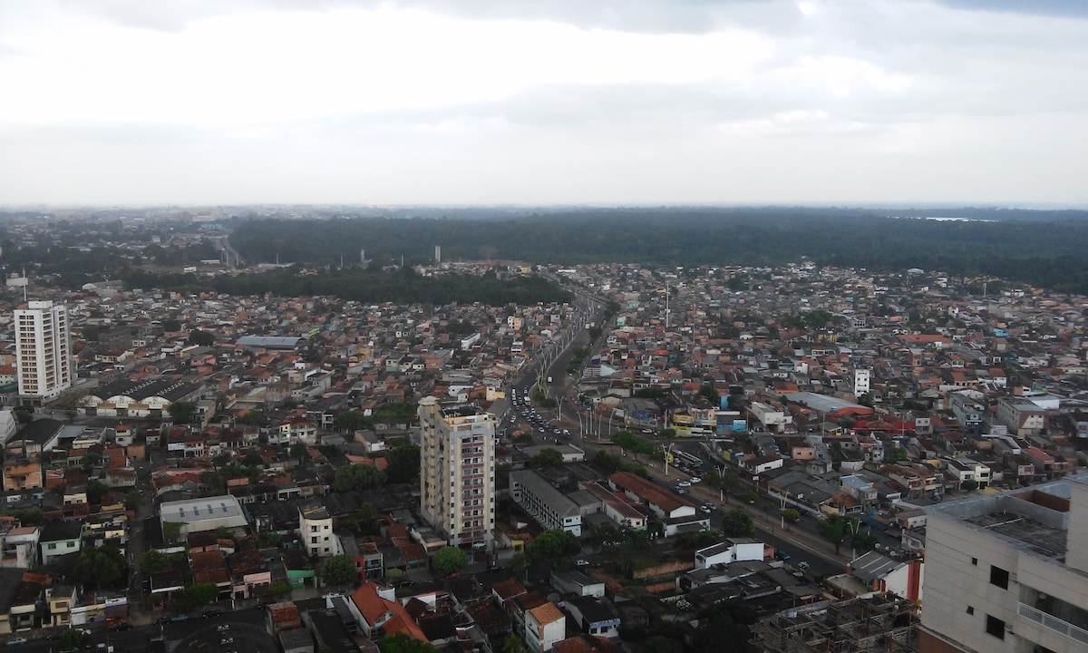 An aerial view captures an expansive urban landscape stretched across the horizon. A mix of residential buildings, roads, and green spaces are visible, highlighting the connectivity between city and nature under a cloudy sky.