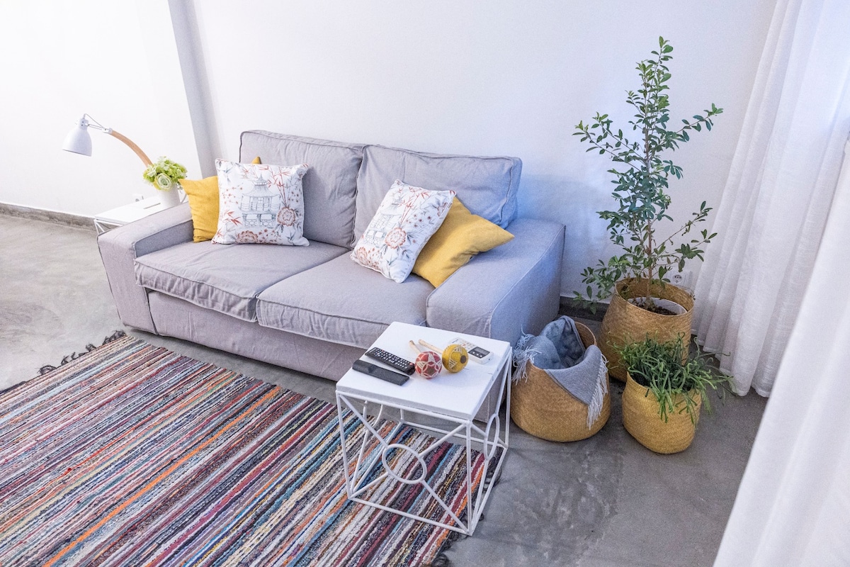 A light-colored couch is complemented by patterned pillows in warm tones. A small coffee table holds various books and decorative items. Potted plants and woven baskets add greenery to the space, while a textured area rug provides warmth underfoot.